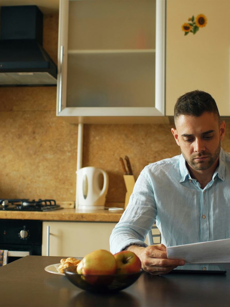 Homem sentado à mesa da cozinha, lendo um documento ao lado de uma xícara de café.